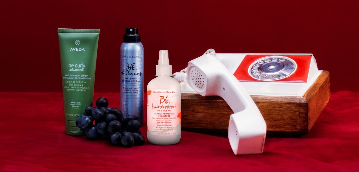 Aveda groene tube krulversterker, Bumble and bumble blauwe aerosol textuur spray, Bumble and bumble witte sprayfles, en zwarte druiven op een rode ondergrond.