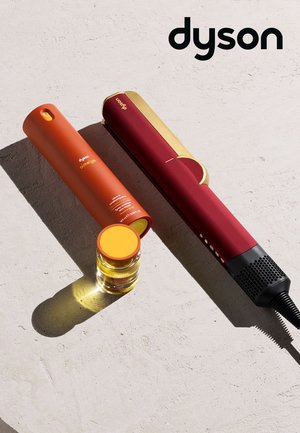 Dyson hårstyling sæt med en rød og guld glattejern, matchende rød hårbehandlingsspray og lille olieflaske på en lys overflade.