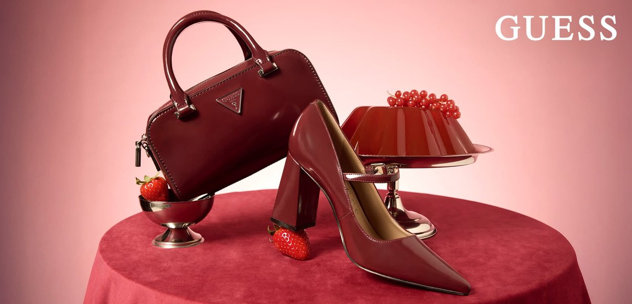 Sac à main bordeaux brillant et chaussure à talon aiguilles pointue présentés avec des fraises et des groseilles rouges sur une surface en velours rouge, logo "GUESS" au-dessus.