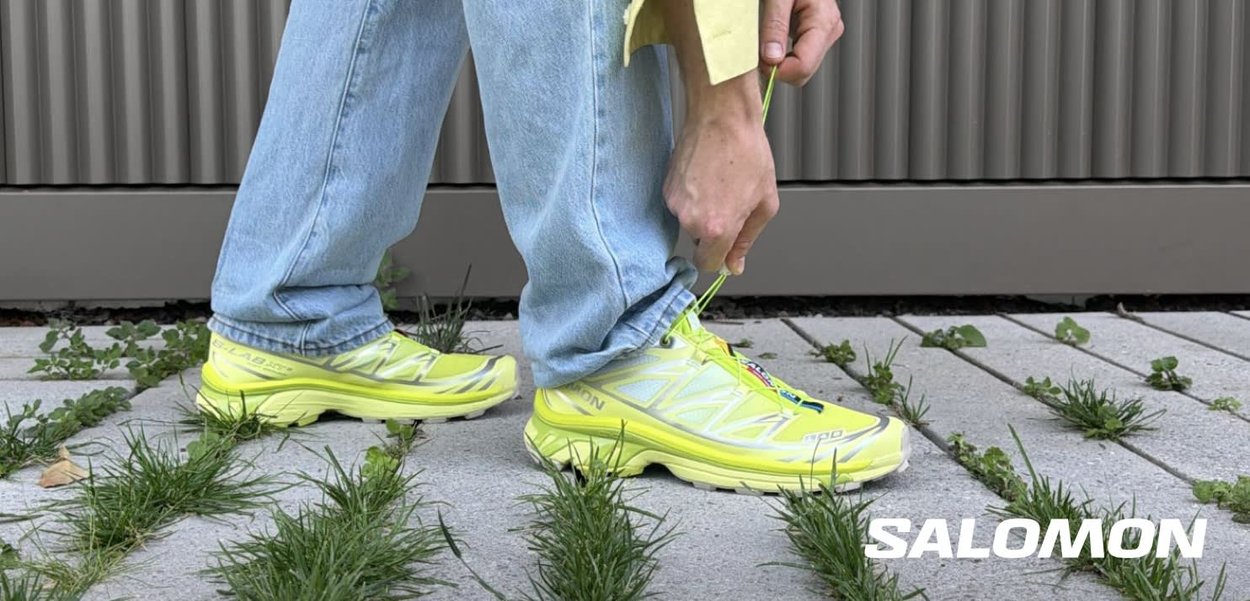 Person i lyseblå jeans trækker op i buksebenets kant og afslører gule Salomon-sneakers på en betonsti med græstuer.