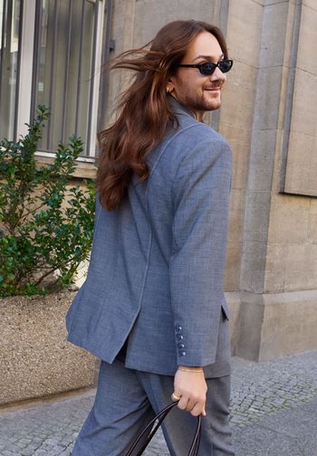 Gray suit with a textured finish, long sleeves, and button accents. Black sunglasses and brown handbag complete the look.