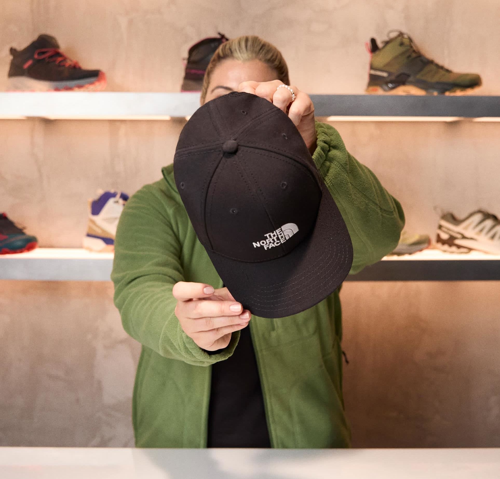 Schwarze Baseballmütze mit einem weißen Logo auf der Vorderseite, gehalten von einer Person in einer grünen Fleecejacke vor einem neutralen Hintergrund mit Schuhen.