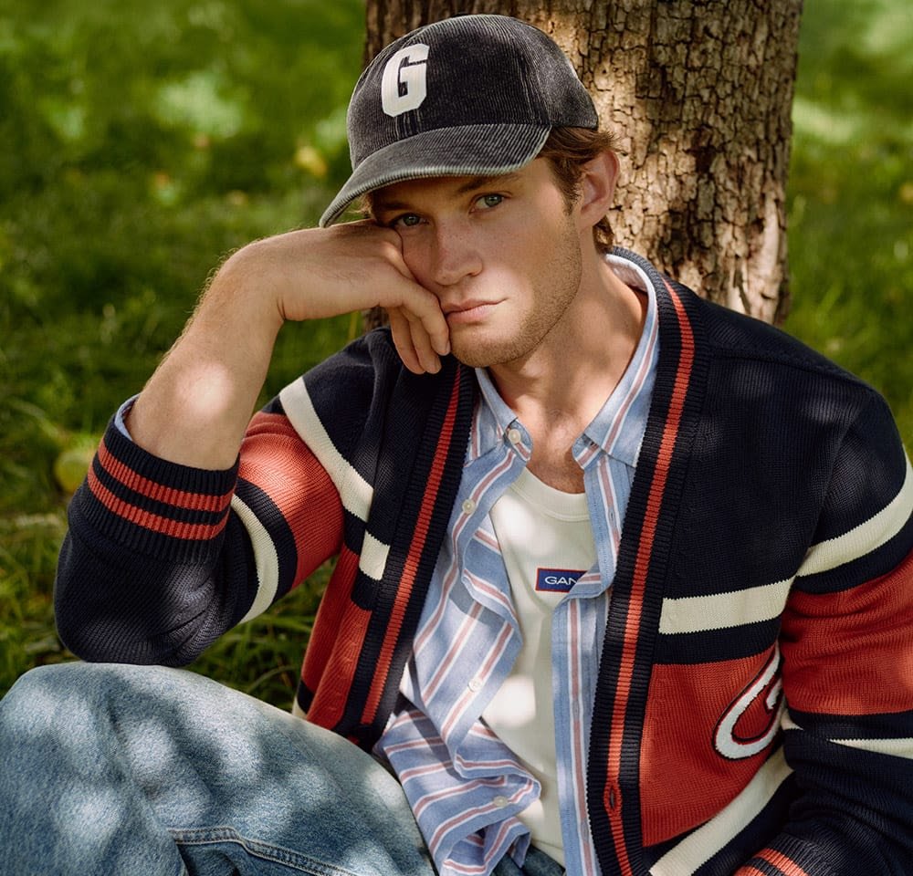 Navy cardigan with red and white stripes, layered over a striped shirt and white tank top, complemented by a black cap featuring a letter "G."
