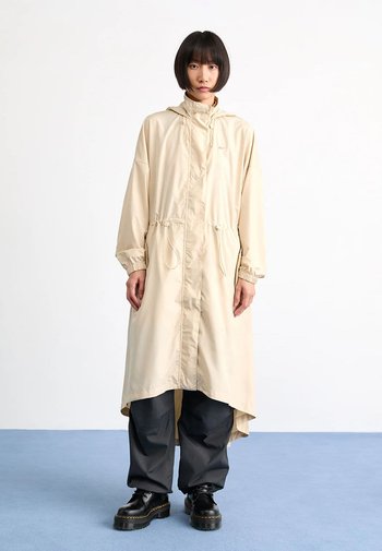 Beige lange jas met een capuchon, aangesloten taille en trekkoord. Draagt over zwarte wijde broek en platform laarzen. Gladde textuur, lichtgewicht materiaal.