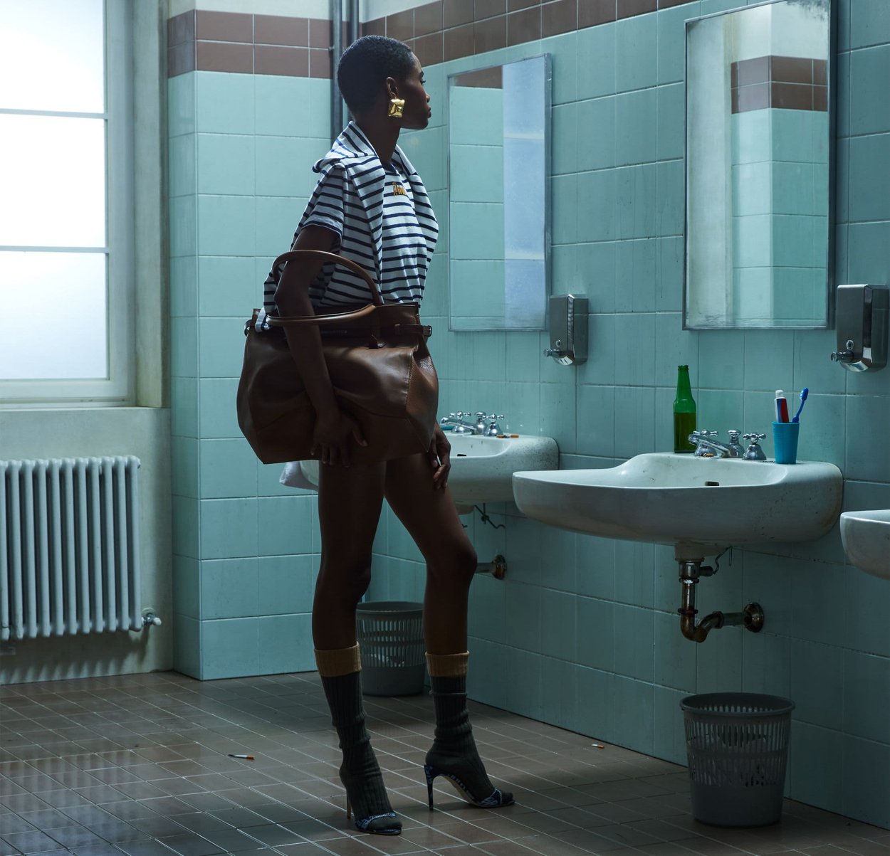 Mujer con camisa de rayas y botas de tacón alto está en un baño público con azulejos, sosteniendo un gran bolso marrón, mirando al espejo sobre los lavabos.