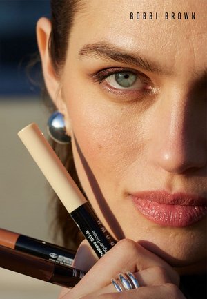 Prodotti di trucco Bobbi Brown tra le mani, con un bastoncino beige chiaro e due bastoncini più scuri. Texture della pelle liscia, orecchini d'argento e labbra naturali.