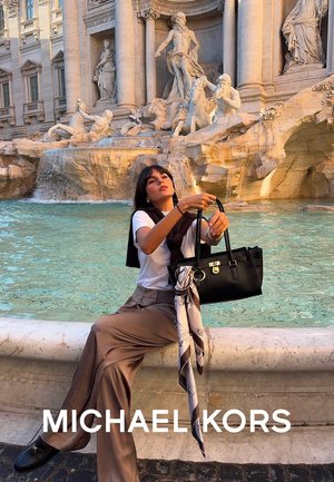 Borsa a tracolla in pelle nera con doppi manici e accessori in argento, accompagnata da una sciarpa fantasia. Pantaloni a gamba larga marroni e camicia bianca.
