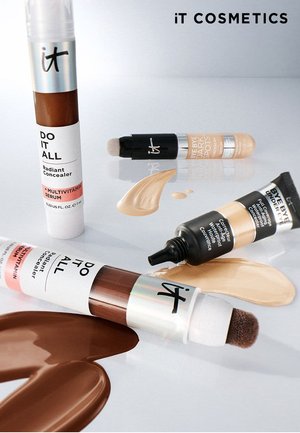 Four tubes of IT Cosmetics concealers with different shades applied on a white surface, featuring applicator tips and product labels.