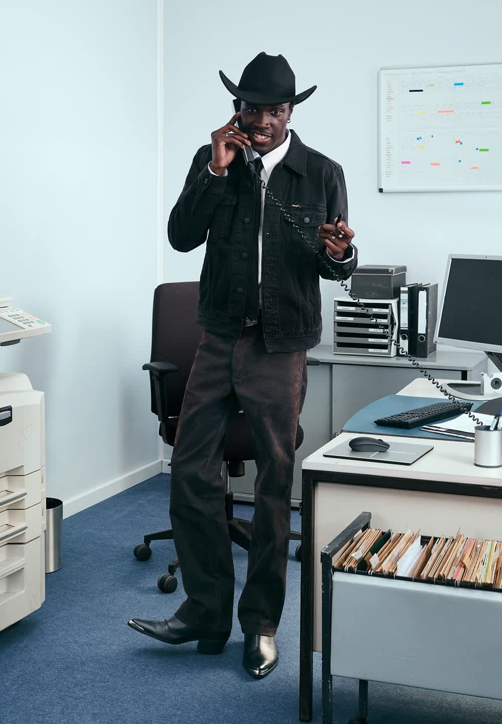 Svarte denimjakke over en hvit skjorte, mørke bukser og svart cowboyhatt. Står ved et skrivebord med en telefon, holder telefonrøret i hånden, i et kontormiljø.
