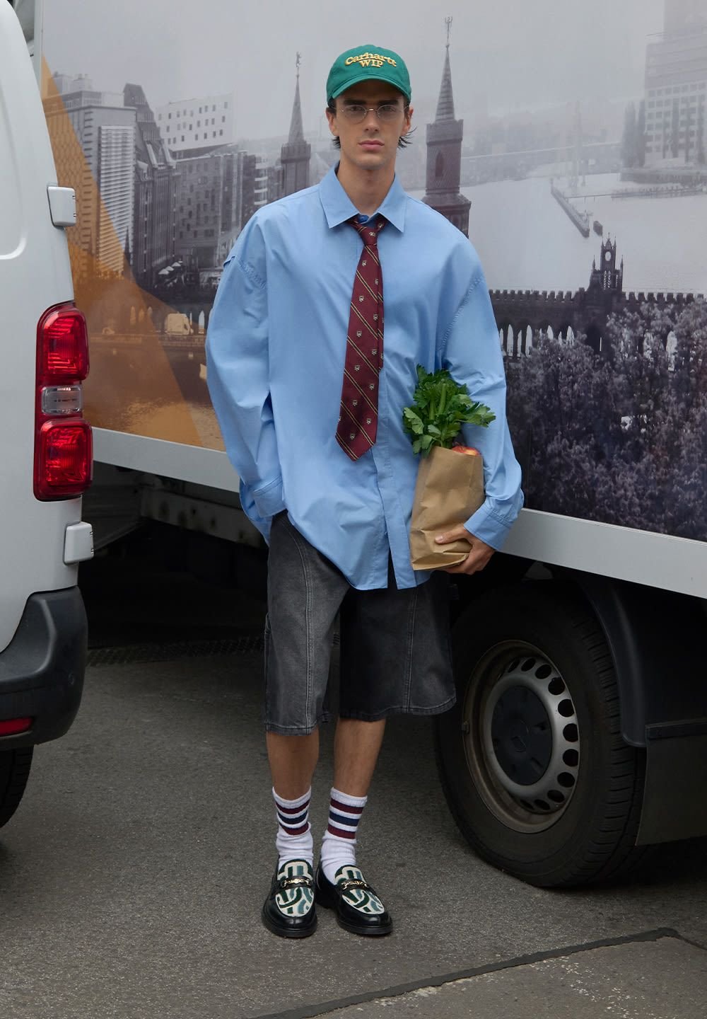 Fiatal férfi kék, nagy méretű ingben, csíkos nyakkendőben, farmer sortban, zöld sapkában és mokaszinban áll, miközben egy papírzacskót tart a boltban vásárolt élelmiszerekkel, közel a tehergépkocsikhoz.