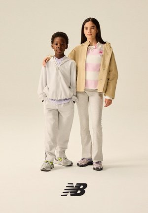 Deux enfants debout côte à côte, vêtus de tenues décontractées New Balance et de baskets, sur un fond beige uni avec le logo NB en dessous.
