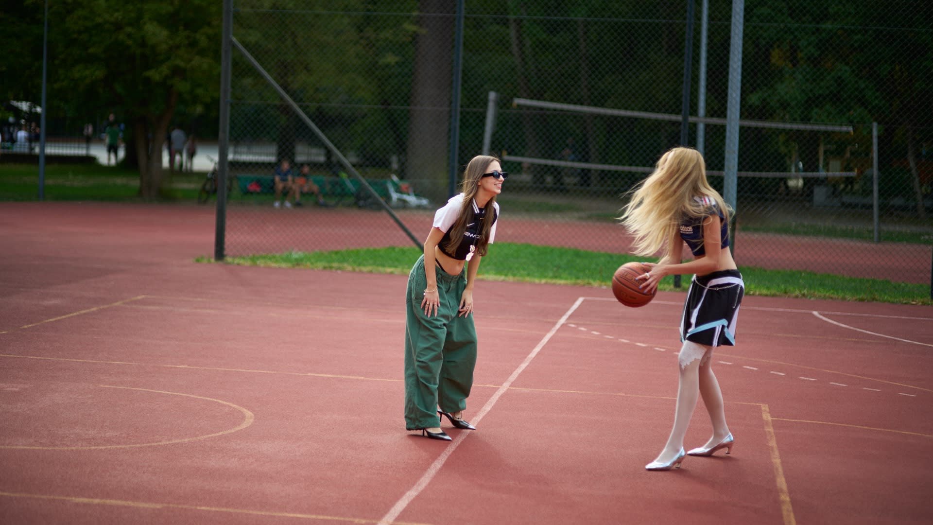 Twee vrouwen op een basketbalveld. De ene draagt een zwarte crop top en groene oversized broek; de andere draagt een blauwe rok en witte hakken terwijl ze met een bal dribbelt.