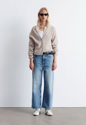 Beige wool cardigan with a relaxed fit, white tank top, light blue wide-leg jeans, and white sneakers with green accents.