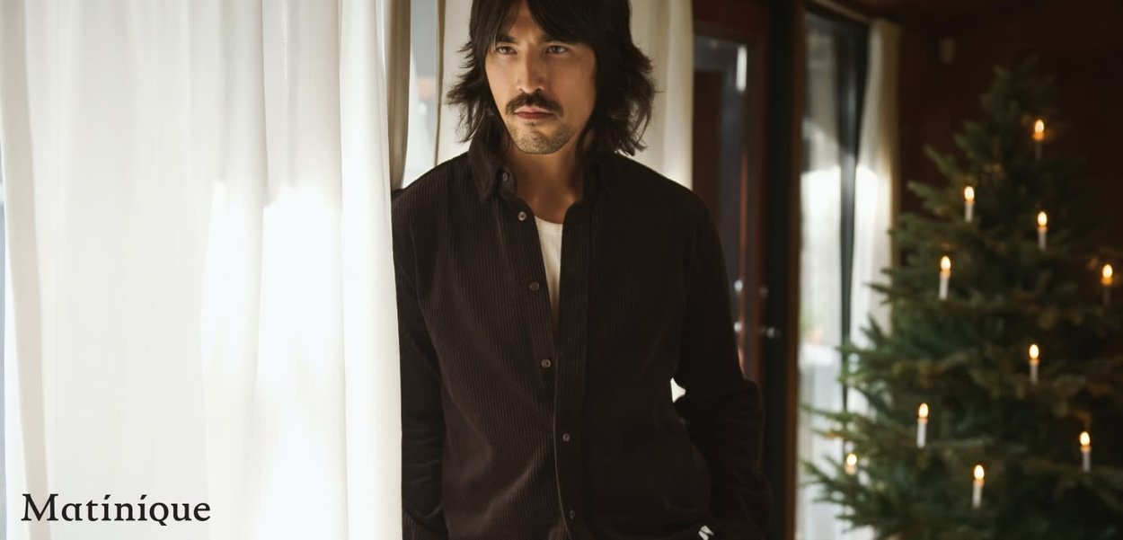 Dunkles Cordhemd mit Knöpfen und lockerer Passform, getragen über einem weißen T-Shirt. Hat lange Haare und steht neben transparenten Vorhängen und einem dekorierten Baum.