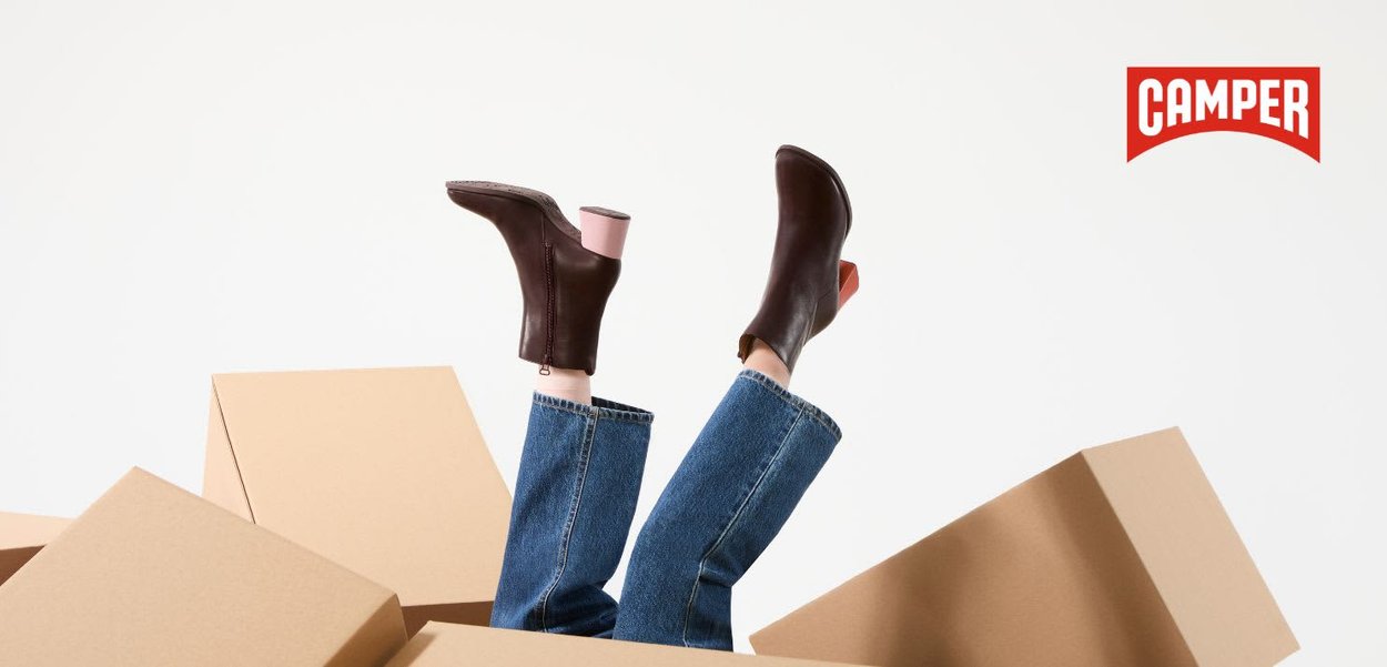Person mit braunen Stiefeletten mit Absatz und blauen Jeans, liegend zwischen verstreuten Pappkartons, oben rechts mit Camper-Logo.