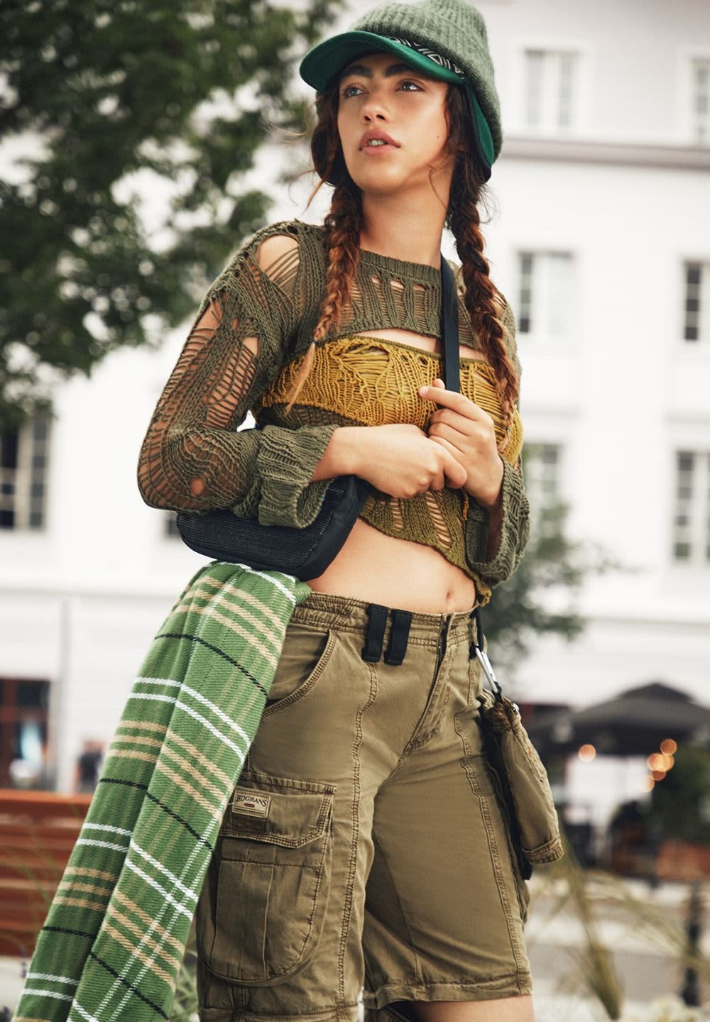 Knit green cropped sweater with cut-out details, yellow underlayer, khaki cargo shorts, green checked scarf, and a black handbag.