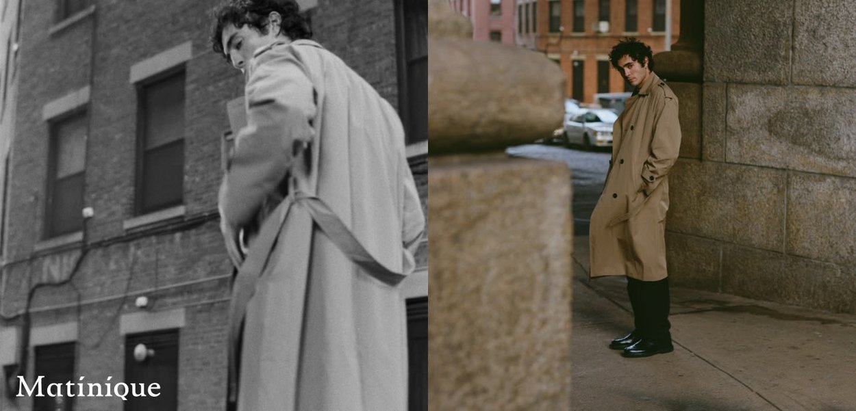 Uomo con trench beige in piedi vicino a edifici urbani, posa con sicurezza con le mani in tasca, sfondo cittadino diurno.