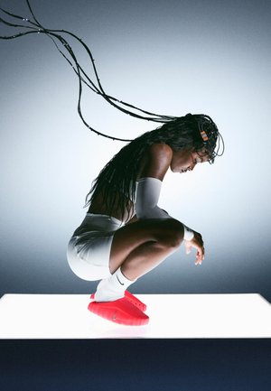 Zapatillas deportivas rojas con parte superior texturizada, pantalones cortos blancos y mangas para brazos. Peinado con alambres. El sujeto está agachado sobre una superficie iluminada contra un fondo gris.