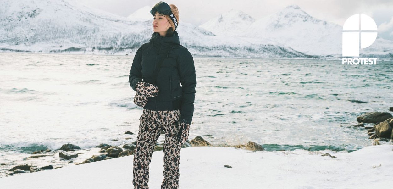 Veste matelassée noire isolante, pantalon de ski imprimé léopard, lunettes de ski et moufles, sur fond de paysage enneigé avec de l'eau et des montagnes.