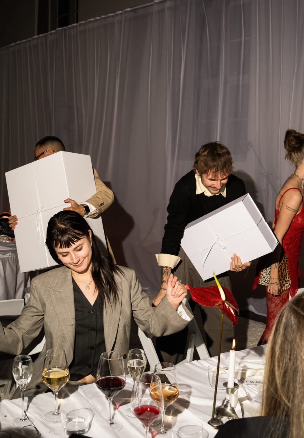 Två personer bär stora vita lådor vid ett bord med olika glas vin. En mörkröd blomma och ett ljus finns också med.