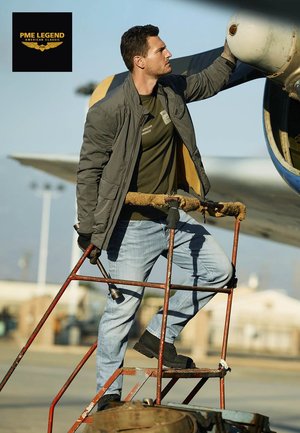 Mann in grüner Jacke und Jeans inspiziert einen Flugzeugpropeller, steht auf einer abgenutzten roten Metallleiter mit Werkzeugen in der Nähe eines Flugplatzes.