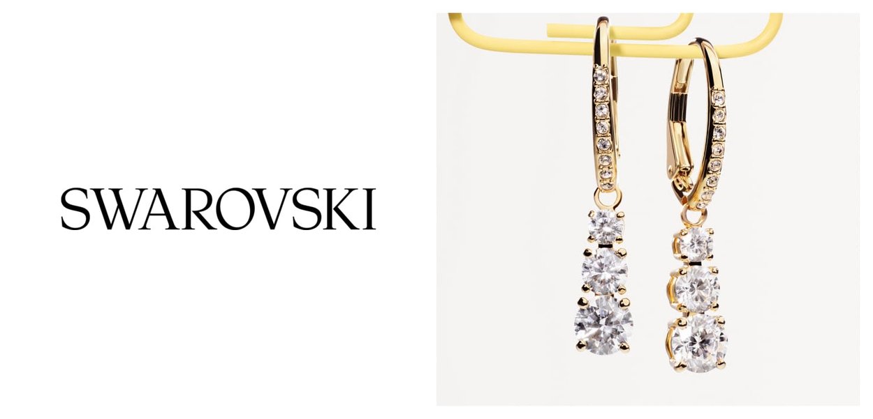 Orecchini a goccia dorati con tre cristalli trasparenti di dimensioni crescenti, appesi a morsetti con chiusura a leva arricchiti di cristalli, logo del marchio Swarovski accanto.