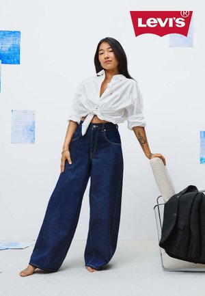 Light blue denim wide-leg jeans with contrast stitching, paired with a knotted white shirt. Model stands barefoot against a white background.