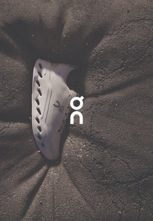 Sneaker bianca con design a basse altezze, caratterizzata da ampie aperture laterali per la ventilazione, tessuto testurizzato e un'estetica minimalista, posizionata su terreno irregolare.