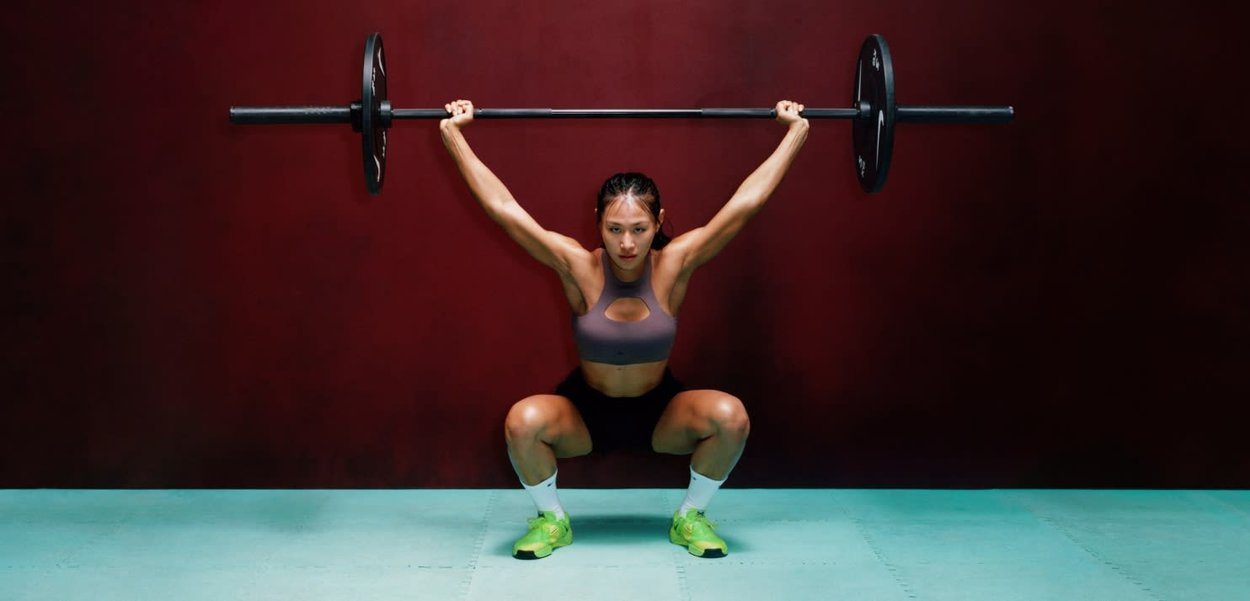 Athlete performing a squat with a barbell, featuring a black bar and weights, wearing a gray crop top and bright green shoes on a blue mat.