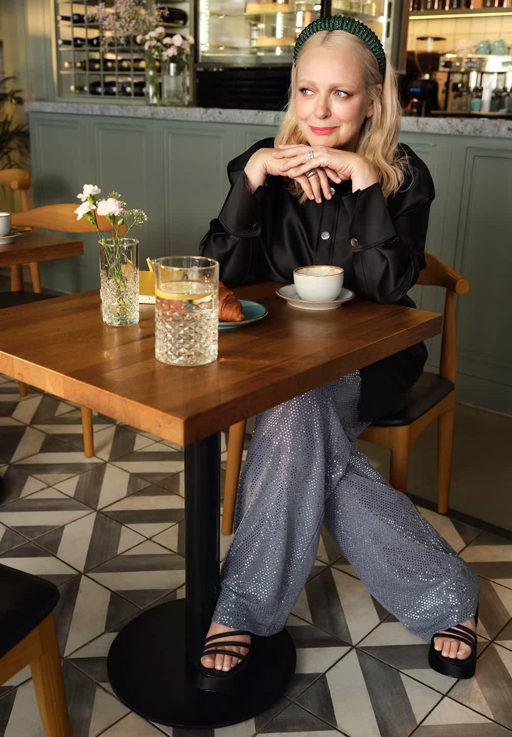Czarna satynowa koszula, błyszczące srebrne spodnie z cekinami, czarne sandały na platformie. Stół z rogalikiem, kawą i kwiatową aranżacją w szklanym wazonie.