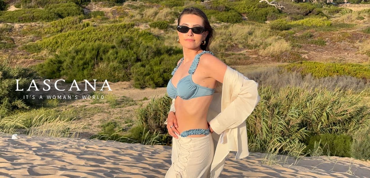 Donna che indossa un top bikini blu e una gonna beige con una giacca beige sulla spalla, in piedi su una spiaggia sabbiosa con vegetazione sullo sfondo.