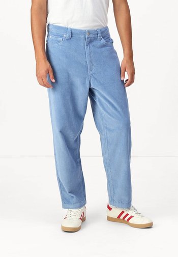 Lichtblauwe corduroy broek met verticale strepen, voorzien van een knoopsluiting, voorzakken en een relaxte pasvorm. G gedragen met witte schoenen.