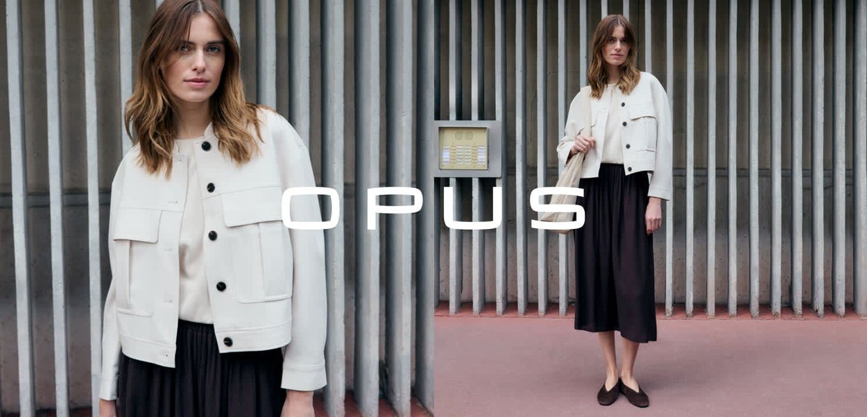 Jeune femme aux cheveux mi-longs portant une veste blanche courte sur une robe midi noire, debout devant un mur à lattes verticales.