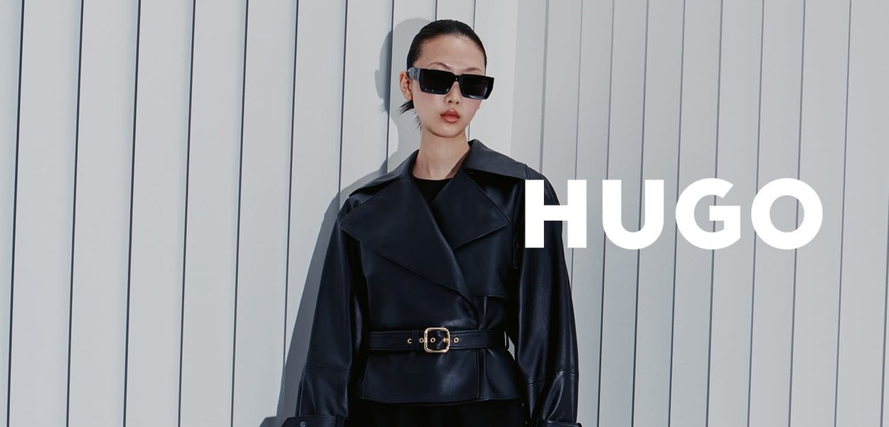 Veste en cuir noire avec larges revers et taille ceinturée, associée à de grandes lunettes de soleil carrées, sur un fond clair.