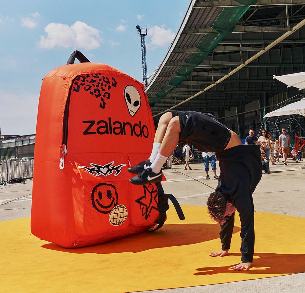 Aufblasbarer orangefarbener Rucksack mit schwarzen Akzenten, der Logos, ein Alien-Grafik und verschiedene Muster zeigt. Steht auf einer gelben Matte im Freien.
