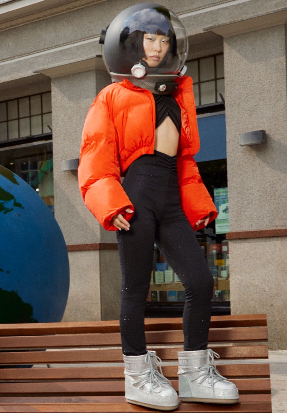Orange puffer jacket with oversized sleeves, black fitted top, black sparkly leggings, and silver lace-up boots, complemented by a space helmet.
