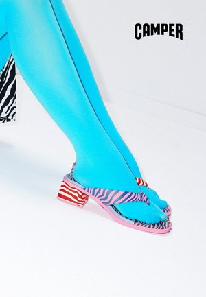 Piernas con medias de color azul brillante que llevan sandalias de tacón a rayas rosas y rojas con diseño en los dedos, logo de Camper sobre fondo blanco.