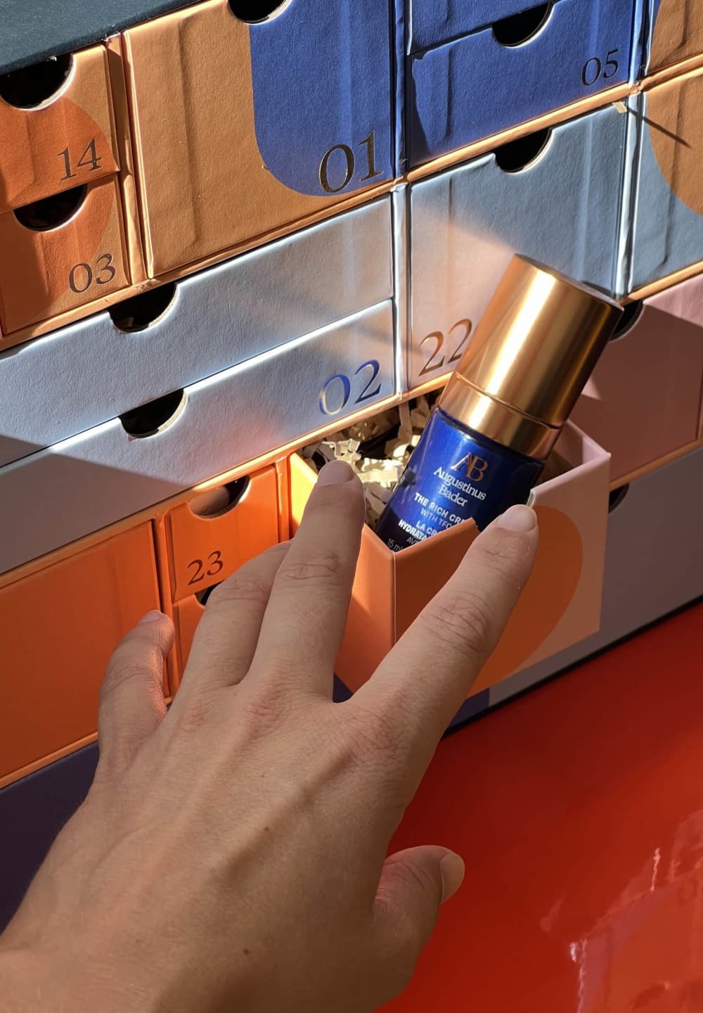 Bouteille en verre bleue avec un bouchon-pompe en or, reposant dans un tiroir orange d'un calendrier de l'Avent multicolore, une main atteignant vers elle.