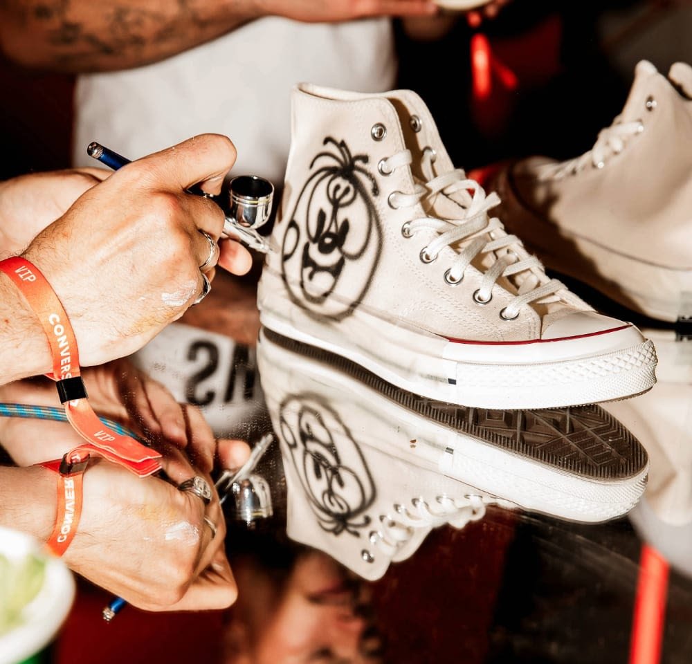 High-top white canvas sneaker with black spray-painted design, red accents on the sole, shown on a reflective surface, hand applying details.