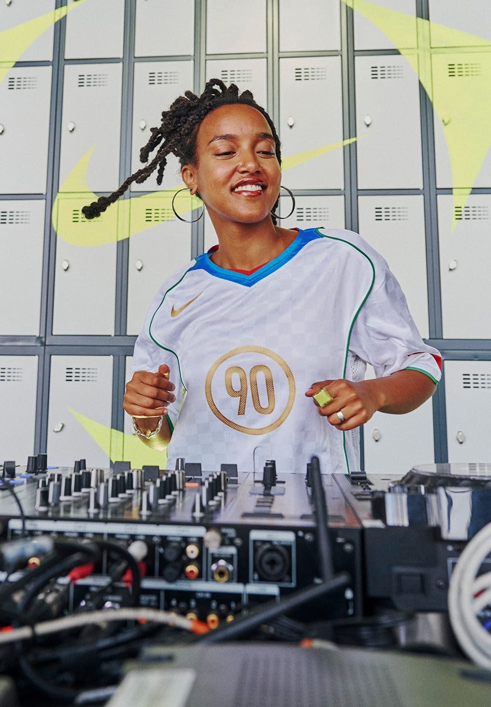 Weißes Nike-Trikot mit blauen und grünen Akzenten, das auf der Vorderseite eine goldene "90" zeigt. DJ-Ausrüstung im Vordergrund; graue Schließfächer im Hintergrund.