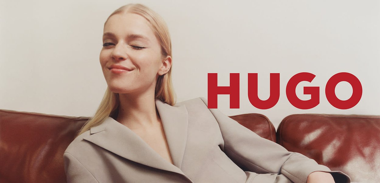 Blazer beige avec de larges revers, texture lisse, porté par une personne sur un canapé en cuir marron. Le mot "HUGO" en lettres rouges audacieuses est affiché.