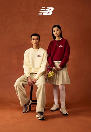 Zwei junge Erwachsene in New Balance Sweatshirts und Sneakern posieren vor einem braunen Hintergrund, einer sitzt auf einem Hocker, der andere hält Blumen.