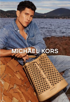 Man in denim shirt en spijkerbroek houdt een grote geweven tan lederen tas vast, zittend op een rotsachtige oever met zee en heuvels op de achtergrond.