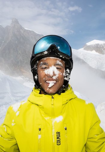 Helle gelbe Skijacke mit Schnee im Gesicht, schwarzem Skihelm und blauen Skibrillen. Berglandschaft im Hintergrund, teilweise bewölkter Himmel.