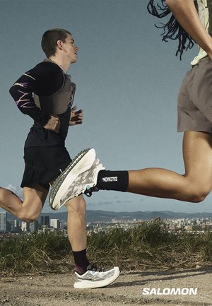 To personer, der løber udendørs på en sti iført Salomon løbesko, med en bysilhuet og klar himmel i baggrunden.