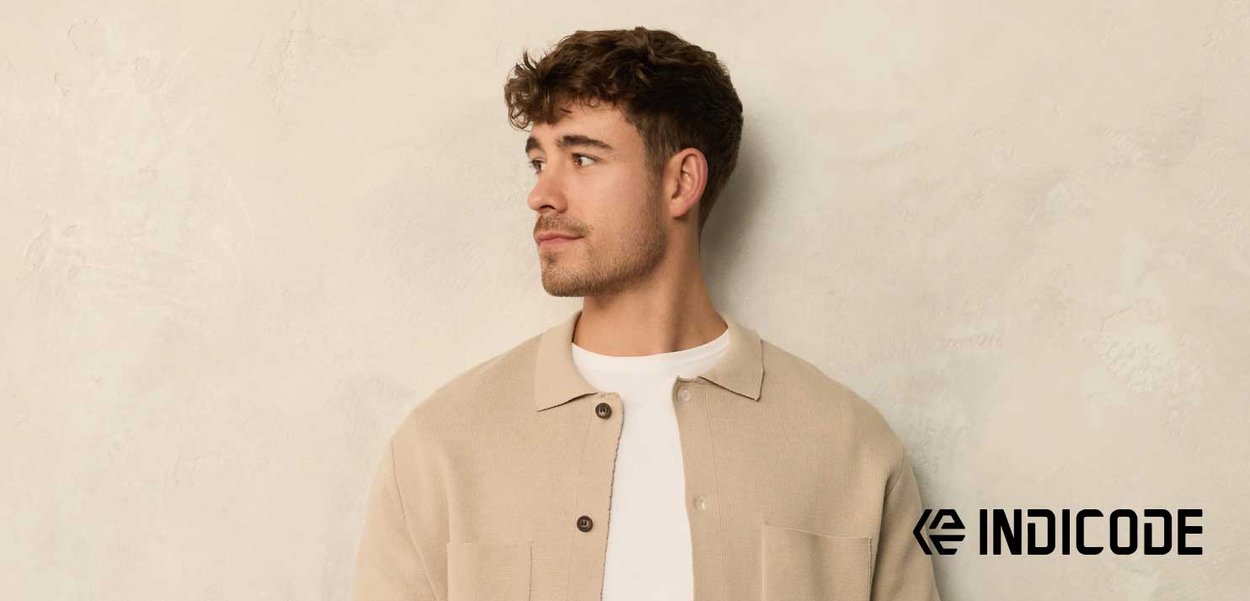 Beige stickad kofta med krage, framknäppning och fickor, bärs över en vit skjorta. Bakgrunden är ljus och texturerad.