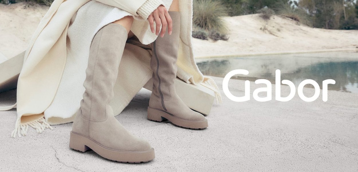 Des bottes en daim beige jusqu'aux genoux avec une semelle plate, présentant des coutures courbes et une finition texturée, positionnées à côté d'une couverture de couleur claire.