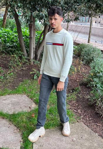 Ljusgrå tröja med röda och blå horisontella ränder, mörkgrå jeans och kraftiga vita sneakers, står i en grön utomhusmiljö.