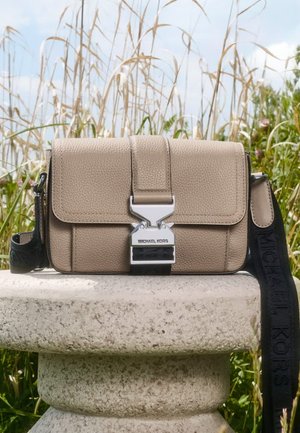 Beige texturerad läderaxelväska med silverdetaljer, framficka med knäppning och ett svart band med varumärkeslogotyp.