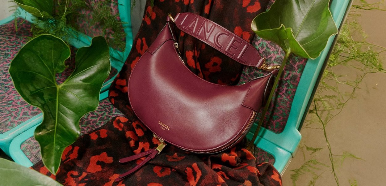 Burgundy leather handbag with a curved shape, embossed 'LANCEL' on the strap, gold hardware, and a zipper, set against a patterned fabric background.